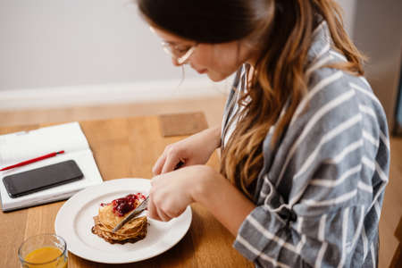 Smiling beautiful woman eating pancakes while having breakfast at home kitchenの写真素材