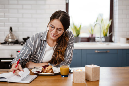 Smiling beautiful woman writing down notes while having breakfast at home kitchenの写真素材