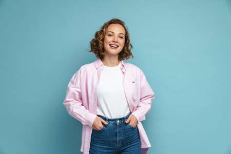 Young ginger woman with wavy hair smiling and looking at camera isolated over blue backgroundの写真素材