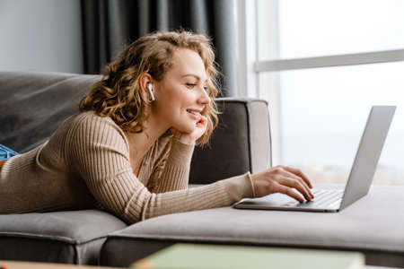 Young woman in earphones using laptop while lying on couch at home