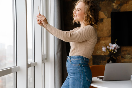 Young blonde woman smiling while taking selfie on mobile phone at home