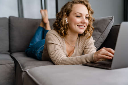 White blonde woman smiling and using laptop while lying on couch at home