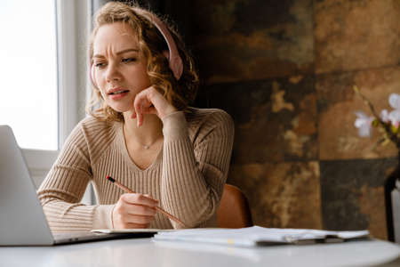Young woman in headphones writing down notes while working with laptop at homeの写真素材