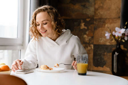 White blonde woman using mobile phone and having breakfast at homeの写真素材