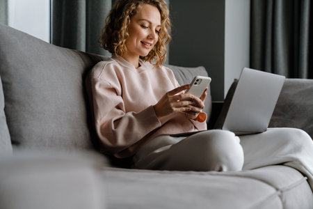 Young white woman using laptop and cellphone while sitting on couch at homeの写真素材