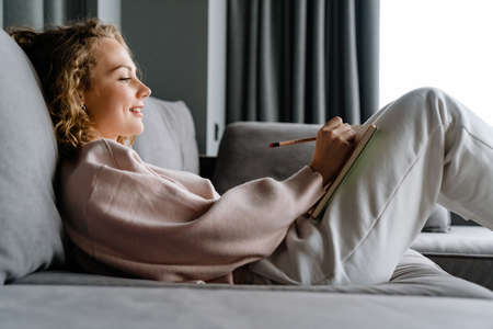Young white woman smiling and writing down notes while lying on couch at homeの写真素材
