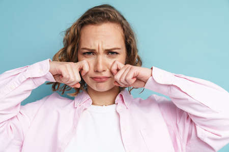Young ginger woman wiping her tears while crying at camera isolated over blue background
