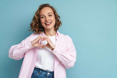 Young ginger woman with wavy hair smiling and showing heart gesture isolated over blue background