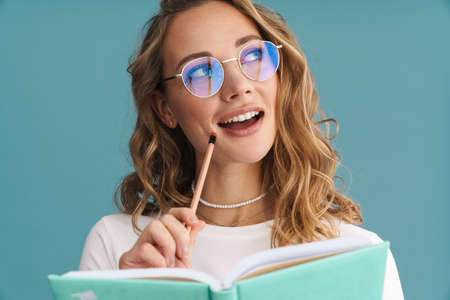 Smiling young woman in glasses holding pencil and book over blue wall background