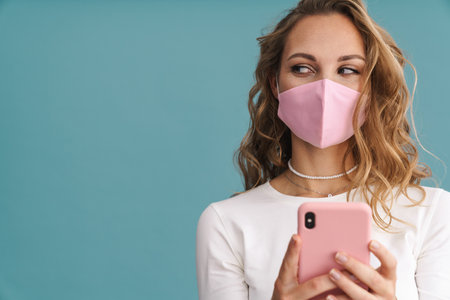 Blonde young woman wearing face mask holding mobile phone over blue wall backgroundの写真素材