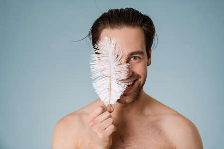 Young smiling shirtless man touching his body with a feather isolated over blue backgroundの写真素材