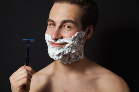 Portrait of a mid aged brunette shirtless man shaving with razor his face over blue wall background