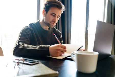 Serious mid aged man working on laptop computer from home, taking notes