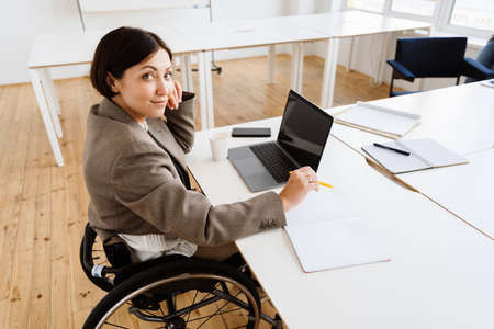 Young woman working with laptop while sitting in wheelchair at officeの写真素材