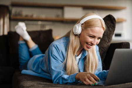 White blonde woman in headphones using laptop while lying on sofa at home