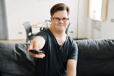 Young man with down syndrome using remote control while sitting on couch at homeの写真素材