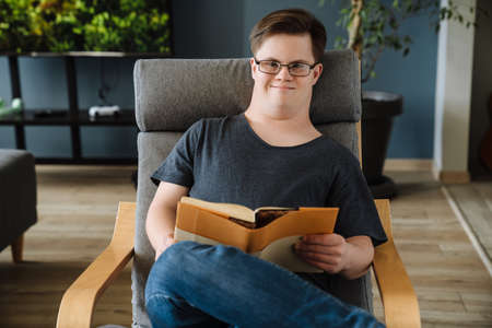 Young man with down syndrome reading book while sitting in armchair at homeの写真素材