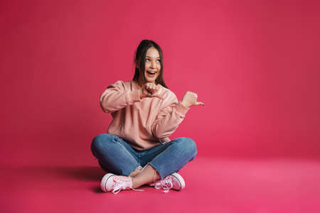 Asian excited woman pointing finger aside while sitting on floor isolated over purple wall
