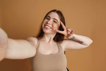 Young ginger woman gesturing peace sign while taking selfie photo isolated over beige wallの写真素材