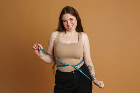 Young ginger woman smiling while posing with measuring tape isolated over beige wallの写真素材