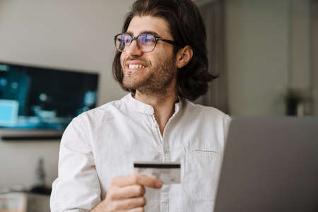 Smiling mid aged brunette turkish man sitting at the table with laptop computer at home holding credit card shopping onlineの写真素材
