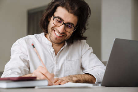 Smiling mid aged brunette turkish man sitting at the table with laptop computer at home writing...