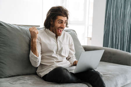 Smiling mid aged brunette turkish man sitting on a couch with laptop computer at home celebratingの写真素材