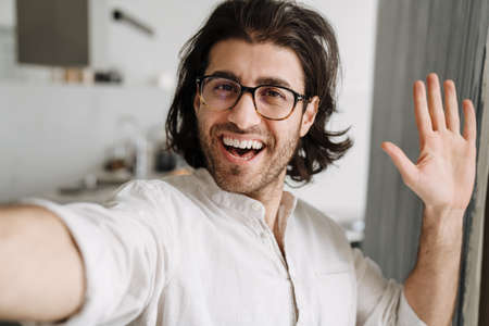 Smiling mid aged turkish brunette man taking selfie standing indoors at home waving handの写真素材