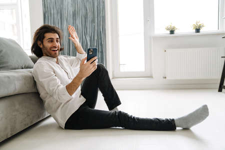 Smiling mid aged turkish man holding mobile phone sitting on a floor in the living room on a video callの写真素材
