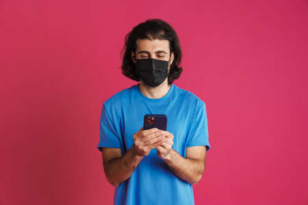 White brunette man in face mask using mobile phone isolated over pink backgroundの写真素材