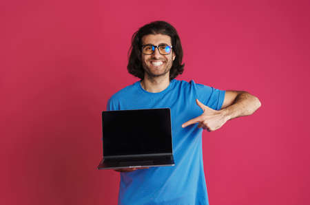 Unshaven smiling man holding and pointing finger at laptop isolated over pink background