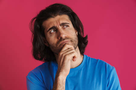 Puzzled brunette man in t-shirt think and looking upward isolated over pink background