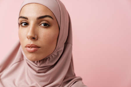 A close-up view of the muslim woman in pink hijab looking at the camera while standing in the pink studioの写真素材