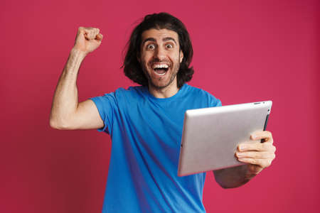White excited man making winner gesture while using tablet computer isolated over pink backgroundの写真素材