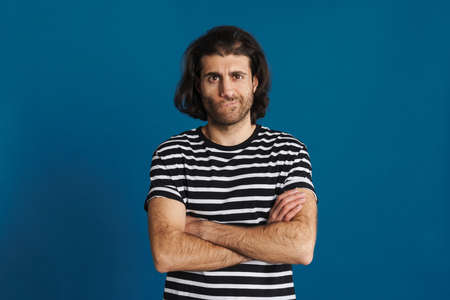 Displeased brunette man in striped t-shirt posing with arms crossed isolated over blue backgroundの写真素材