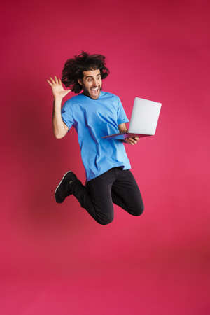 Excited brunette man gesturing and using laptop while jumping isolated over pink background