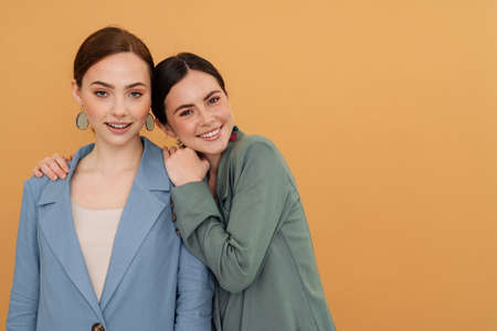 Young two women wearing jackets hugging and smiling at camera isolated over yellow background