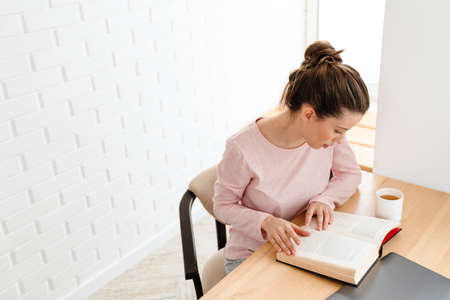 Top view of a young white woman in lounge wear sitting reading book at the table at home
