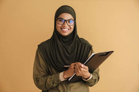Black muslim woman in eyeglasses writing down notes on clipboard isolated over beige backgroundの写真素材