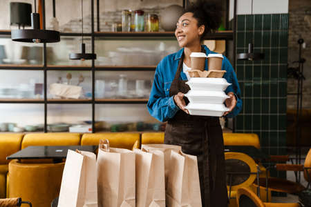 Black waitress in apron holding food boxes while working at cafe indoors