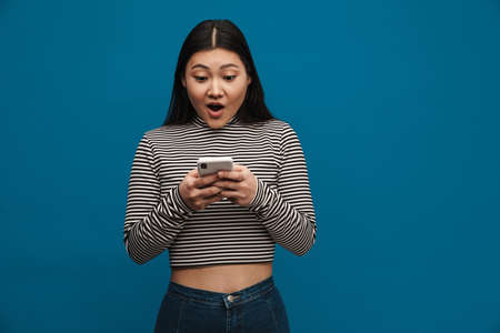 Portrait of an excited casual young asian woman standing over blue wall background looking at mobile...