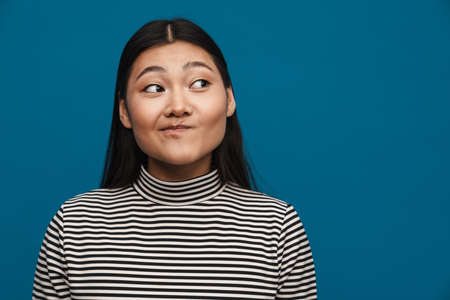 Portrait of a confused casual young asian woman standing over blue wall background looking aside