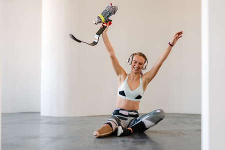 Disabled happy young white sportswoman in headphones with prosthetic leg sitting on a floor in room