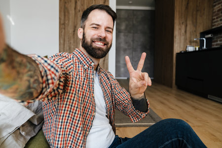 Portrait of a smiling man sitting on the floor and making selfie photo, peace gestureの写真素材