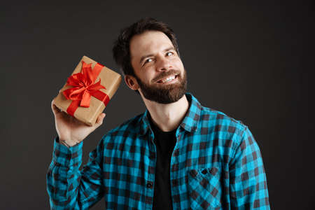 Bearded happy white man smiling while posing with gift box isolated over black backgroundの写真素材