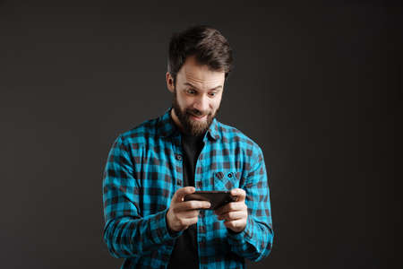 Bearded excited man playing online game on mobile phone isolated over black backgroundの写真素材
