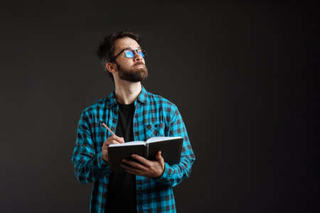 Bearded brunette man in shirt looking upward while writing down notes isolated over black backgroundの写真素材