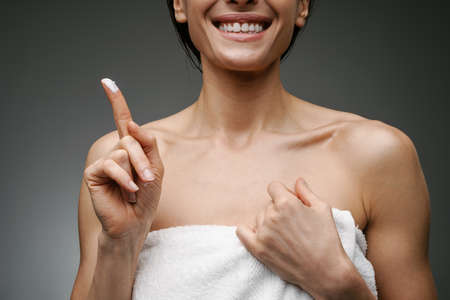 White smiling woman dressed in towel showing facial cream isolated over grey background