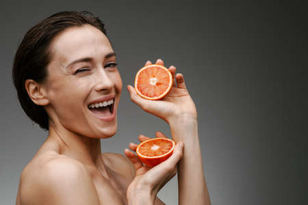 Smiling healthy mid aged topless woman holding grapefruit over gray wall background, winkingの写真素材