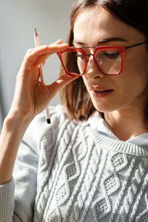 Smiling middle aged woman in sweater holding glasses and pencil indoors